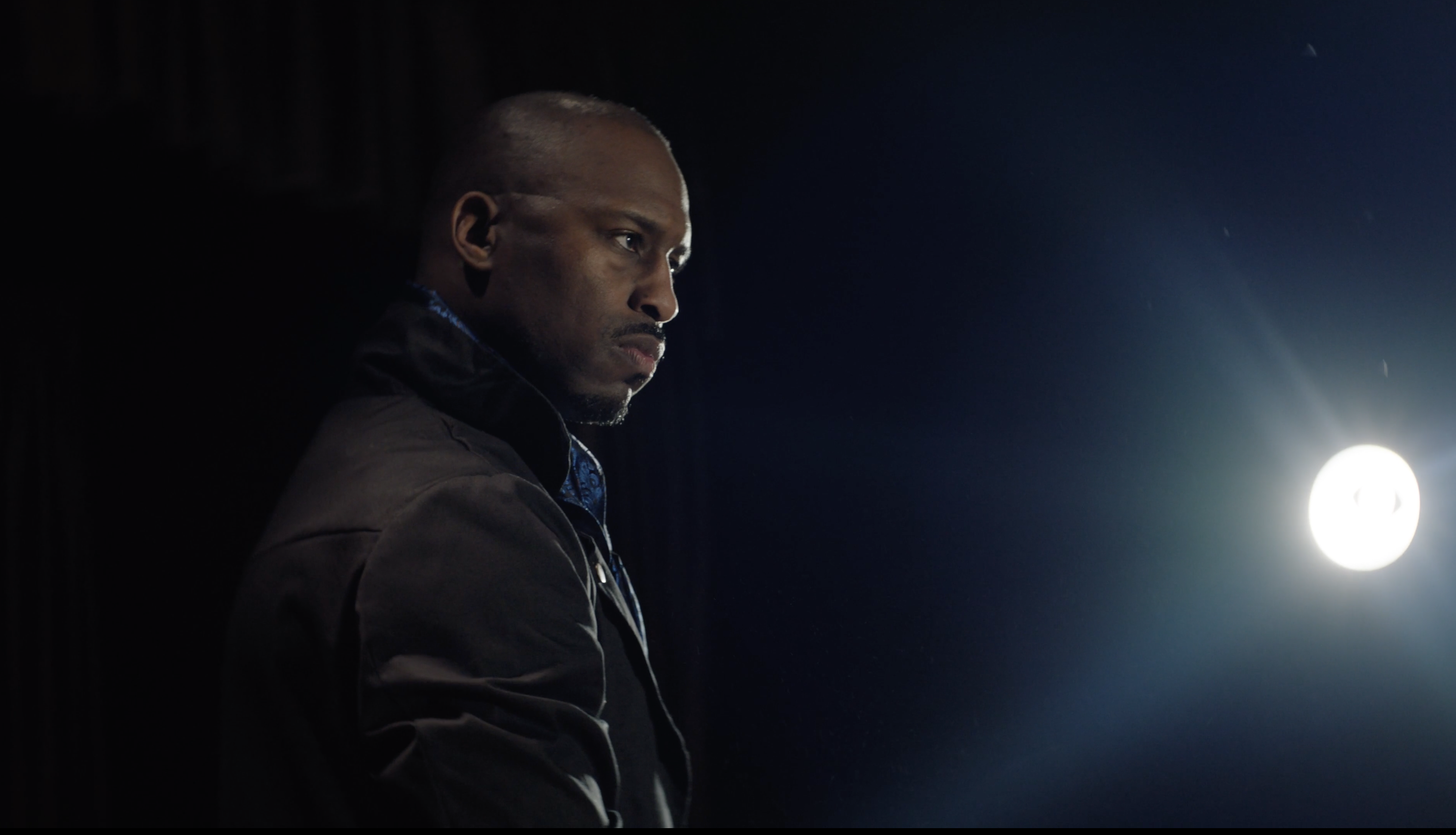 A man in a black coat strikes a pose on stage as a light is shown in the distance.